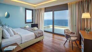 A hotel room with a bed, a desk, a chair, and a large window offering a view of the ocean. - Sparkling ocean views with heated pool (Rodhia)