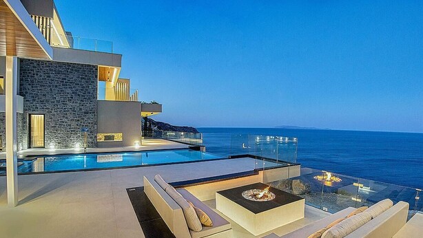 Un área de piscina exterior moderna con barandilla de vidrio, una chimenea y una zona de estar con cojines.