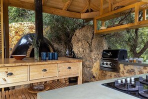 A rustic outdoor kitchen with a stone wall, wooden cabinetry, and a built-in grill.