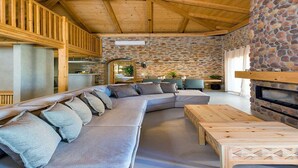 A modern living room with a stone wall, wooden ceiling beams, a large sectional sofa, and a fireplace. - Unique 4 bedroom villa with total privacy (Agios Nikolaos)