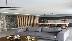 A modern living room with a grey sofa, a wooden coffee table, and a kitchen area in the background. - Seafront 5 bedroom villa (Agia Pelagia)