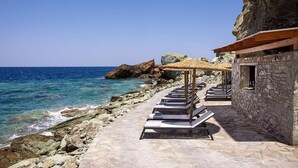 A coastal hotel with stone walls, a stone pathway, and lounge chairs by the sea. - Seafront 5 bedroom villa (Agia Pelagia)