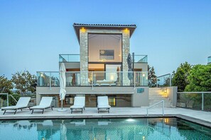 A modern poolside area with lounge chairs, a glass balustrade, and a stone facade building with a sign. - Seafront 4 bedroom villa (Tersanas)