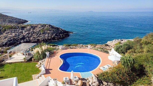 Ein Pool mit gewölbter Kante, umgeben von einer Sonnenterrasse mit Liegestühlen und einer Pergola.