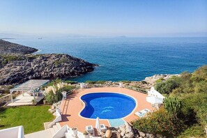 Una piscina con borde curvo, rodeada por una terraza con sillones reclinables y una pérgola.