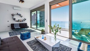 Uma sala de estar moderna com uma janela ampla que oferece vista para o mar, uma mesa de centro branca, um sofá preto e uma estante circular original.