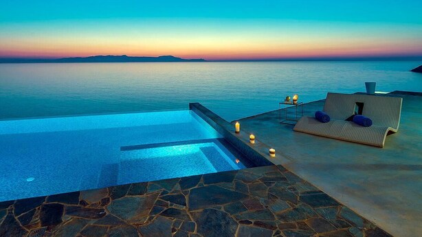 Uma piscina infinita com vista para o mar e um espaço de estar com almofadas e velas.
