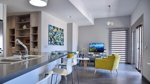 Eine moderne Küche mit Barbereich, Esstisch und Fernseher in einem Wohnbereich mit großem Fenster und Jalousien.