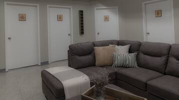 A living room with a brown leather sofa, a small ottoman, and a coffee table with a remote control. There are three white doors with labels indicating different rooms.
