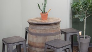 Een rustieke bar met een houten vat als centrale tafel, omringd door twee krukken. Een potplant staat op het vat, en er hangt een kunstwerk aan de muur.