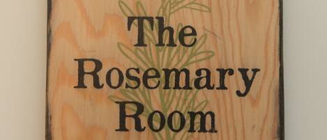 A square wooden sign with the text "The Rosemary Room" and a green rosemary plant illustration.