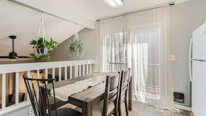 A dining area with a wooden table, chairs, a white railing, hanging plants, and a window with sheer curtains. - Charming Bungalow - Longmont- Nearby Lyons Pet Friendly fenced yard (Longmont)