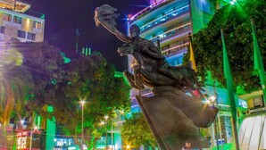 Vista nocturna de una plaza pública con una estatua destacada, edificios modernos y un área pavimentada con gente.