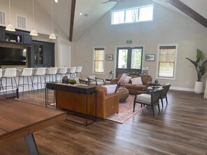 Uma sala de estar ampla, com piso de madeira, um sofá grande, uma mesa de centro e uma área de cozinha com banquetas.