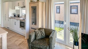 A modern kitchen with white cabinets, a built-in oven, and a coffee maker. There is a grey sofa with a cushion and a potted plant on the balcony.