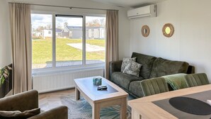 A living room with a sofa, a coffee table, and a dining table. There are two windows with curtains, a radiator, and an air conditioning unit on the wall.