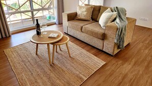 Una sala de estar acogedora con un sofá beige, una mesita redonda con una botella y copas, y un ventanal con vistas a la vegetación.