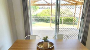 Un área de comedor con una mesa de madera y sillas blancas, una planta en maceta y vistas a un patio exterior a través de una puerta de cristal.