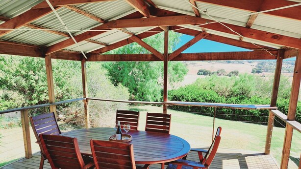Un patio techado al aire libre con una mesa redonda y cuatro sillas de madera, rodeado por una terraza de madera y barandilla de metal.