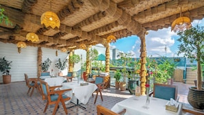 An outdoor dining area with wooden tables and chairs, covered by a thatched roof, and surrounded by potted plants. - Riad Diamond Of Marrakech (Marrakech)