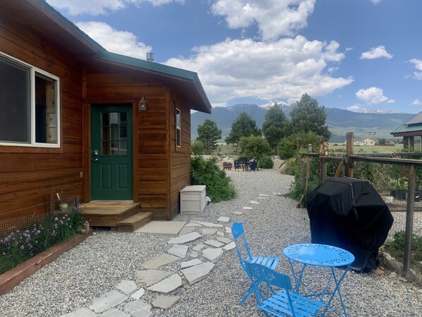 Una cabaña de madera con una puerta verde, un sendero de piedra que lleva a una zona de estar al aire libre azul y una parrilla para barbacoa.