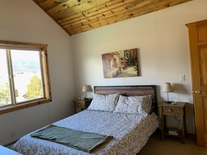 A bedroom with a wooden ceiling, a bed with patterned bedding, a painting on the wall, and a window with a view.