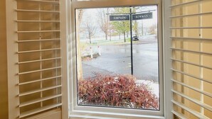 Une fenêtre avec des stores blancs, donnant sur une rue où l’on peut voir les enseignes de « Simpson » et de « Bowman », et un petit jardin agrémenté de feuillage rouge.
