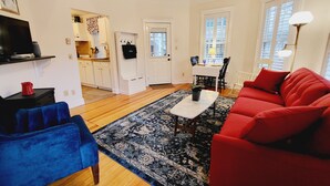 Un salon avec un canapé rouge, un fauteuil bleu et une table basse. Au fond, on aperçoit une cuisine avec des meubles blancs et un coin repas comprenant une table et des chaises.
