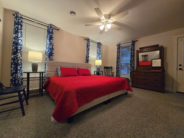 A bedroom with a bed, a desk, a chair, a dresser, a ceiling fan, and windows with curtains.