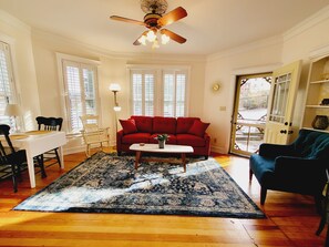 A living room with a red sofa, a white coffee table, and a blue armchair. There is a ceiling fan, a floor lamp, and a dining table with chairs.