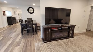 Un soggiorno con TV a schermo piatto, pavimento in legno, tavolo da pranzo con sedie, mobile TV scuro e tappeto decorato.