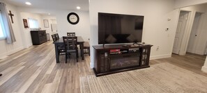 A living room with a flat-screen TV, wooden flooring, a dining table with chairs, a dark entertainment unit, and a patterned rug. - 75 Acre Nature Home 100yd Shooting Range, Axe Throwing, Trails, Fire Pit, & More (Saint Charles)
