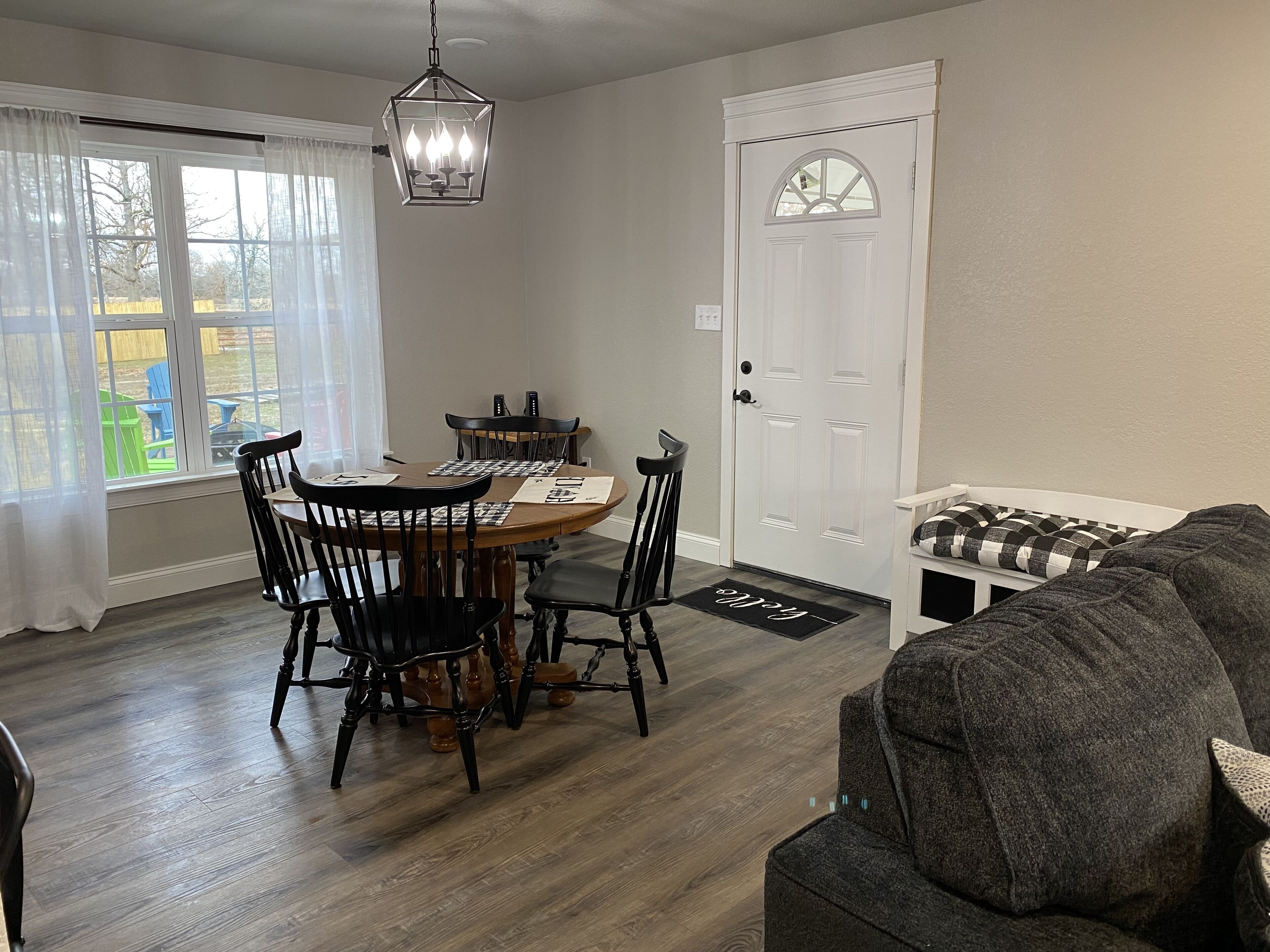 Un coin repas avec une table et des chaises en bois, un lustre, une porte blanche avec une fenêtre et un canapé gris.