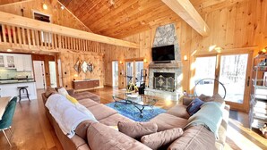 A spacious living room with a large sectional sofa, a stone fireplace, and a kitchen area visible through an open door. - Your Vermont Mountain Haven Pet Friendly home with Hot tub! (Winhall)