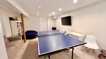 Een kamer met een tafeltennistafel, een witte bank, een relax fauteuil, een witte stoel en een televisie aan de muur.