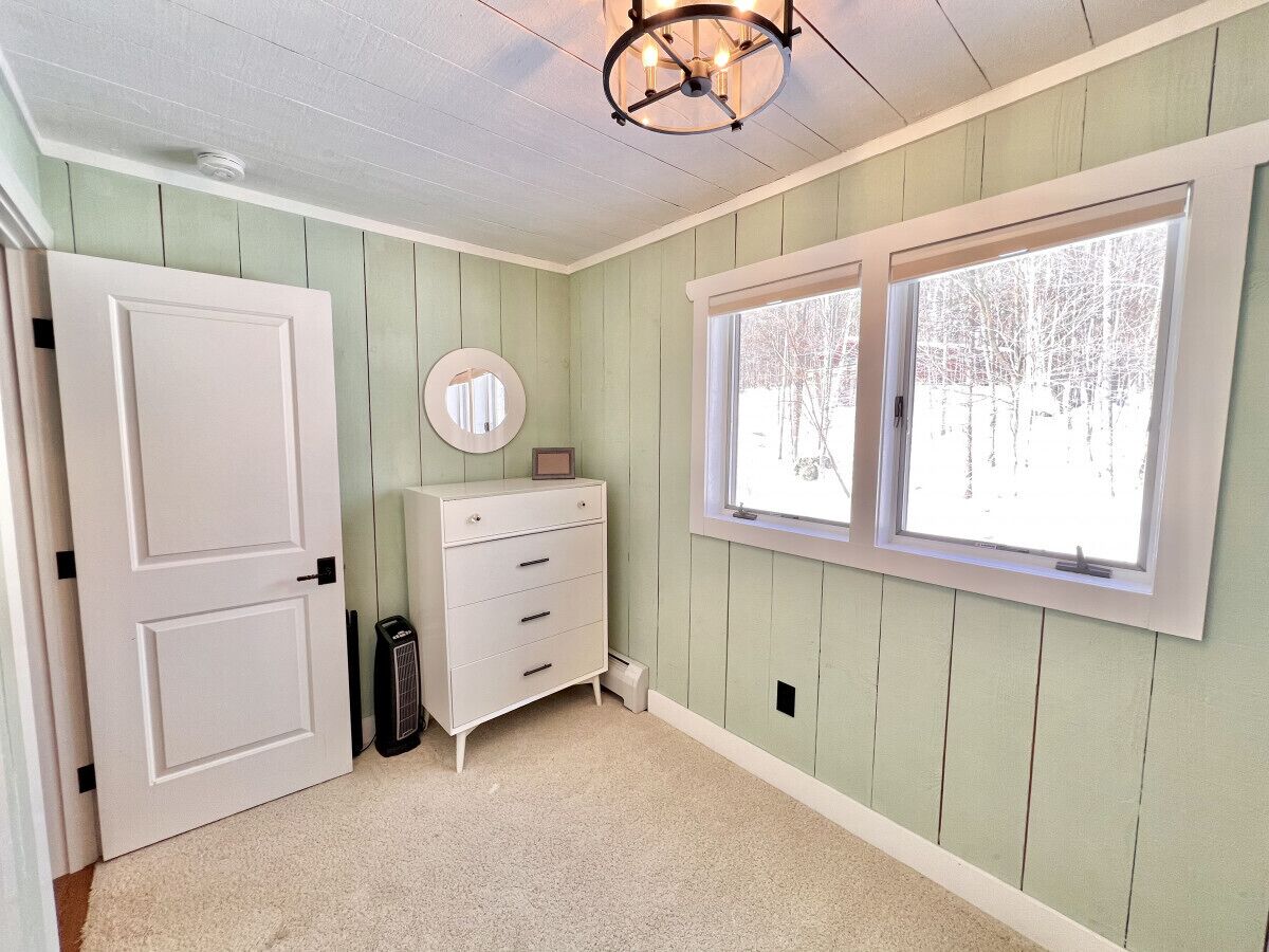 A room with a white door, a dresser with drawers, a round mirror, and a window with a view of trees.