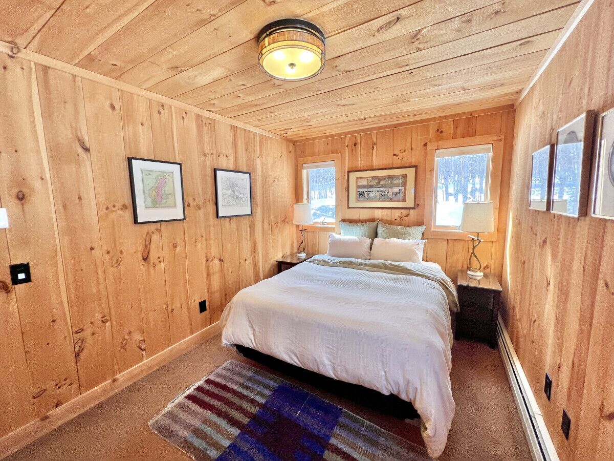 A wooden-paneled bedroom with a bed, framed pictures, a nightstand, and a window with a view of trees.