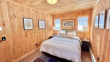 A wooden-paneled bedroom with a bed, framed pictures, a nightstand, and a window with a view of trees.