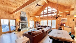A cozy cabin interior with a wooden interior, a fireplace, a sofa, a dining table, and large windows. - Your Vermont Mountain Haven Pet Friendly home with Hot tub! (Winhall)