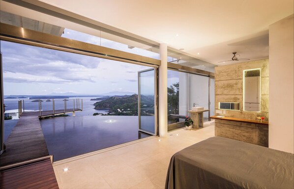 A modern bedroom with a large window overlooking a body of water and hills.