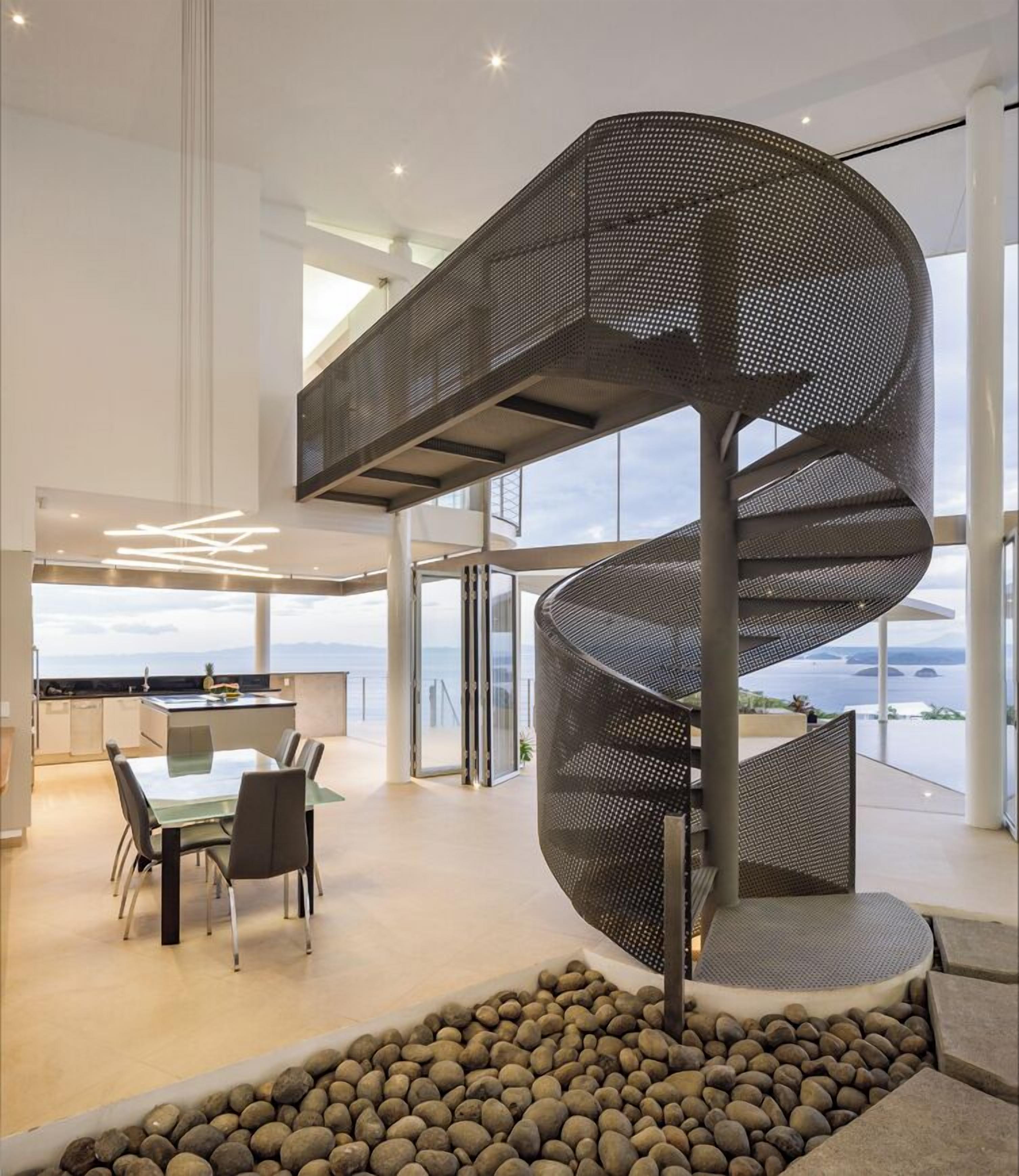 Een modern interieur met een spiltrap, een eethoek met een glazen tafel, en uitzicht op zee.