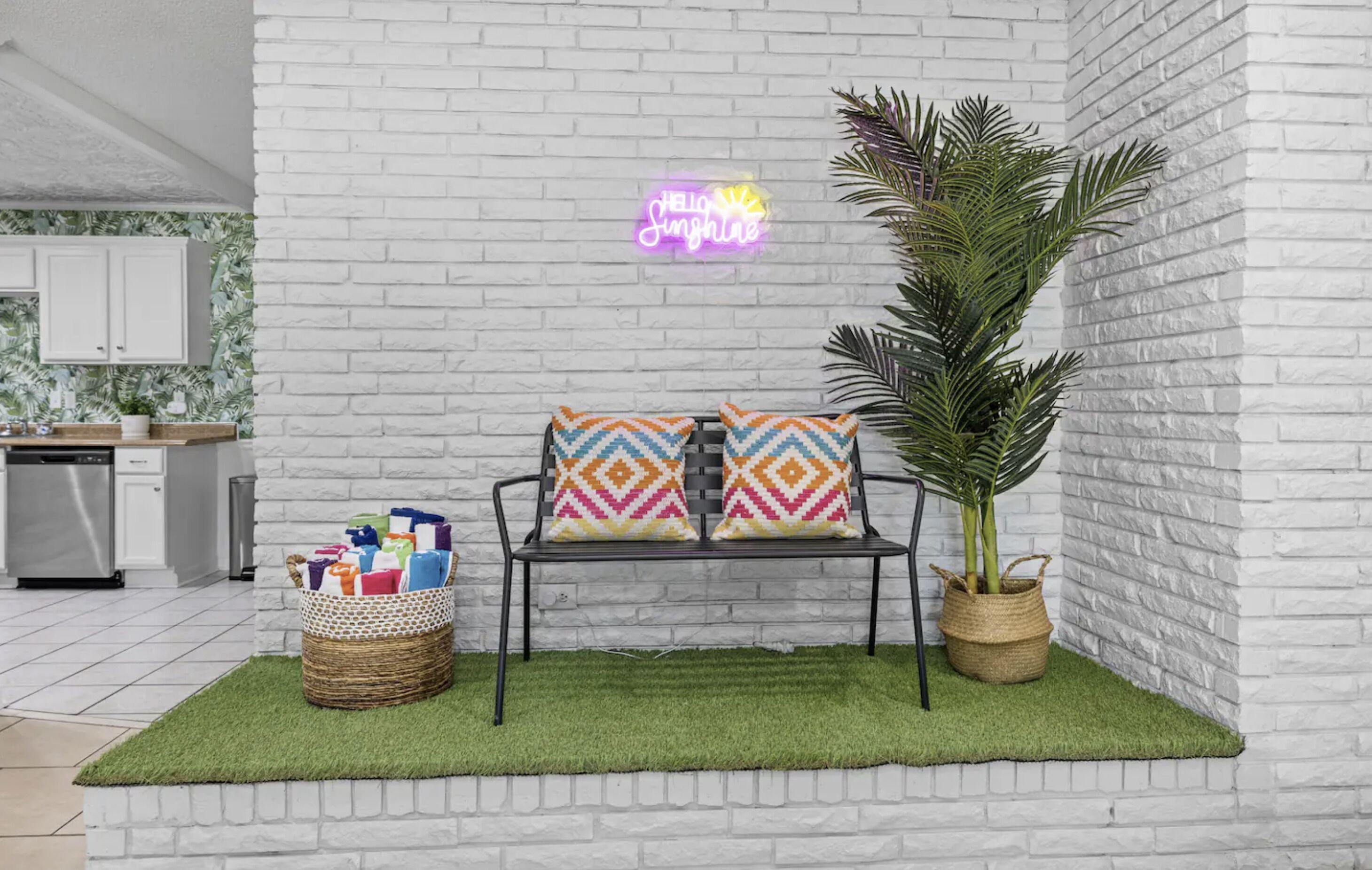 A modern kitchen with white cabinets, a neon sign, a bench with patterned cushions, and a potted plant.