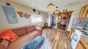 A cozy living room with a sofa, a coffee table, and a rug. There is a kitchen area with wooden cabinets and a stove. A map of the United States is on the wall, and a clock is also visible.