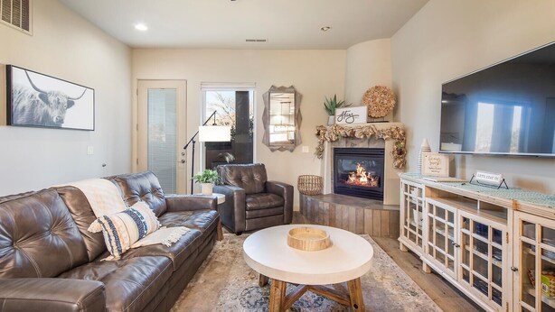 A living room with a leather sofa, a round coffee table, a fireplace, a TV, and a cabinet with glass doors. - Spacious, Hidden Gem at Point Vista! (St George)