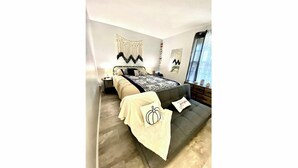 Un dormitorio con cama, mesitas de noche, una ventana con cortinas y un macramé colgado en la pared.