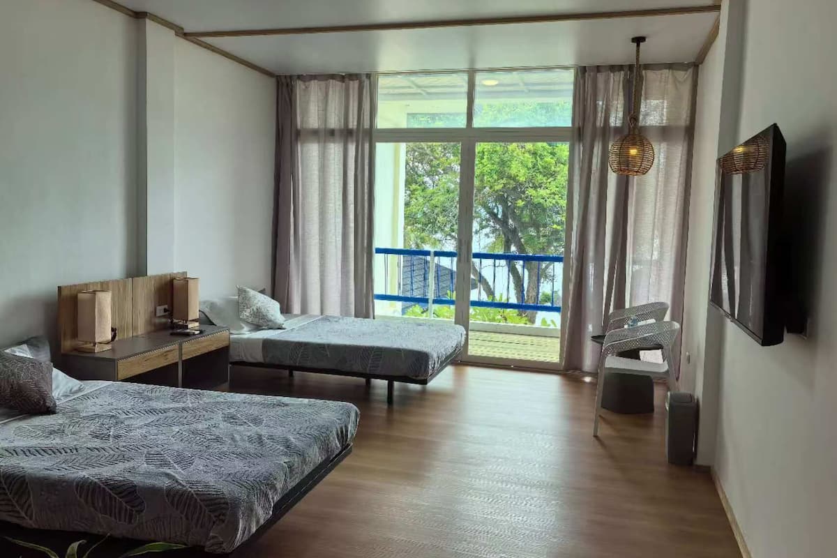 Ein Zimmer mit zwei Einzelbetten, einem kleinen Tisch mit Lampe, einem an der Wand befestigten Fernseher und einem Balkon mit blauer Brüstung.