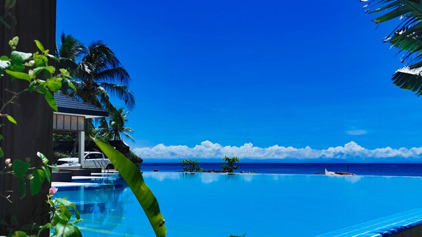 A infinity pool with a clear view of the ocean and a small boat in the distance. - Blue Joys Marine Sports Resort (San Juan)