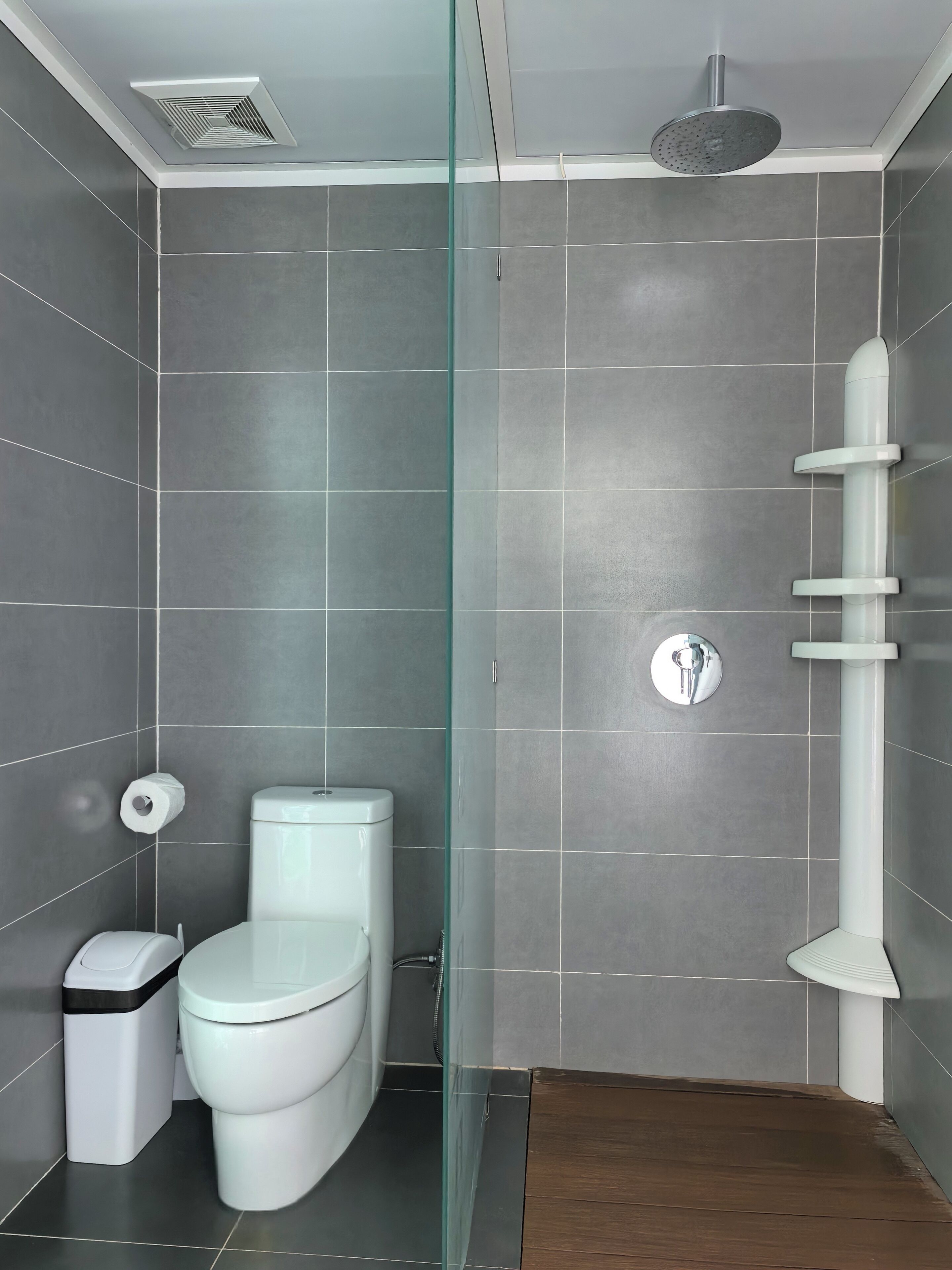 A modern bathroom with grey tiles, a white toilet, a glass shower enclosure, and a wooden floor.