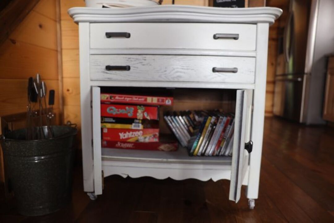 A white cabinet with two drawers and a shelf containing books and a box labeled "Yachtzee."