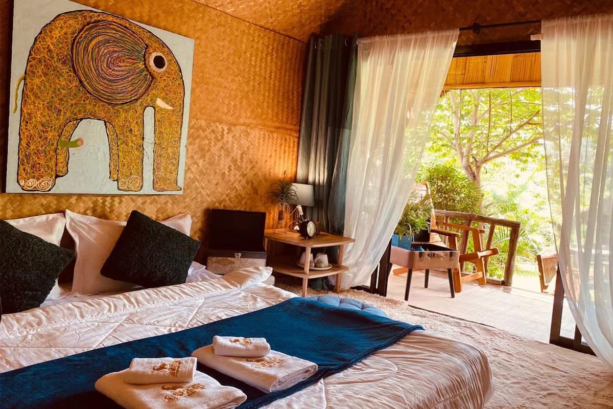 Ein Schlafzimmer mit einem großen Bett, einem Schreibtisch, einem Stuhl und Blick auf Bäume durch transparente Vorhänge.